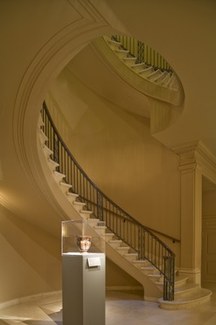 A photograph of ISAW's foyer, showing the large spiral staircase going up and an ancient ceramic object in a display case in front of it.