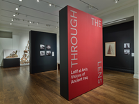 A large gallery room. In the foreground a standing wall partition reads “Through the Lens, Latif Al Ani’s Visions of Ancient Iraq.” Behind this is a second wall partition with mounted photographs. The gallery walls have mounted photographs, paintings, and