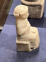 Photo of a small statue of a seated, robed person in a museum display case.