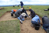 Photo of a group of nine archaeological team members excavating square grids in a grassy field with mountains in the background