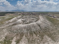 Aerial photo of a mound, with settlement site on the far side, and mountains in the distance.