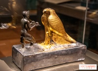 Photo of museum display case containing statue depicting kneeling king offering small globe-shaped vessels to large falcon deity.