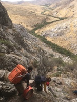 Two people with camping gear traverse rocky terrain with the view from the Tuttybulaq 2 cave which includes many brown colored hills and a river like green stream running through the middle.