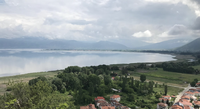 View across Lake Orestiada toward the Neolithic lakeshore site of Dispilio, with green fields and red-roofed houses in the foreground and mountains rising in the distance under a cloudy sky.