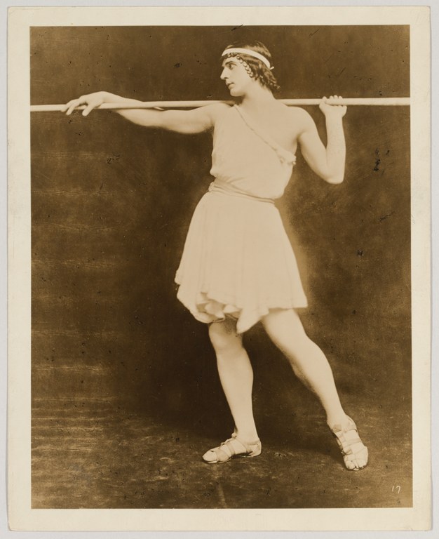 Photograph of a man wearing a short, sleeveless tunic that is tied at the waist and that leaves his knees bare. His head is turned to the left, with one arm resting on a shoulder-height bar and the other hand gripping it in his fist.