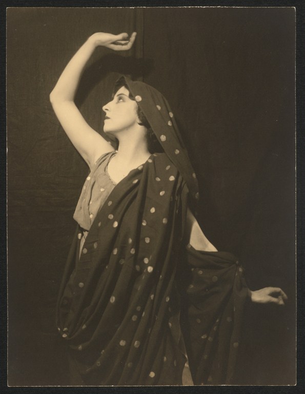 Photograph of a woman wearing a sleeveless dress and a long, shawl over her head and draped over one shoulder and arm. She raises the other hand above her head as she gazes up and to the side.