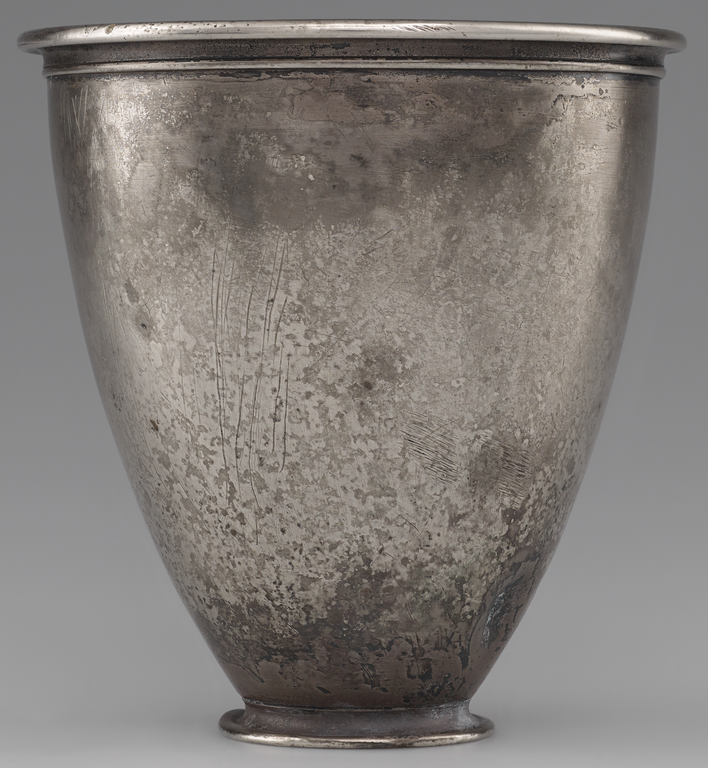 Photograph from the side of a silver beaker, narrow at the base and widening to a simple, protruding rim at the top. No figurative decoration is visible.