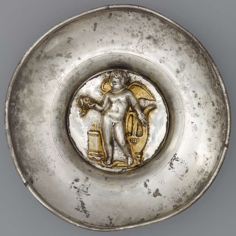 Photograph of a silver bowl with a large, central medallion in silver and gold depicting nude cupid with wings and a stringed musical instrument