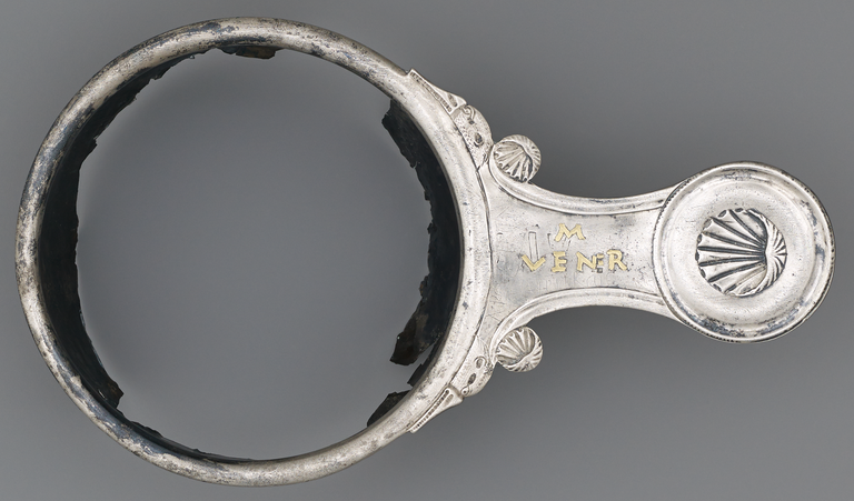 Photograph from above of a silver, long-handled bowl from which the body of the bowl is missing. On the handle there is a silver seashell and some Latin letters in gold.