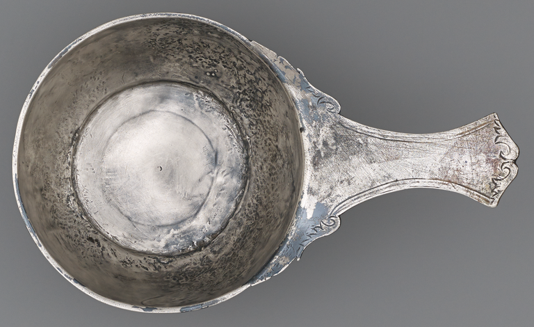 Photograph of a silver, long-handled bowl from above. The only decoration is a simple, incised border on the handle.