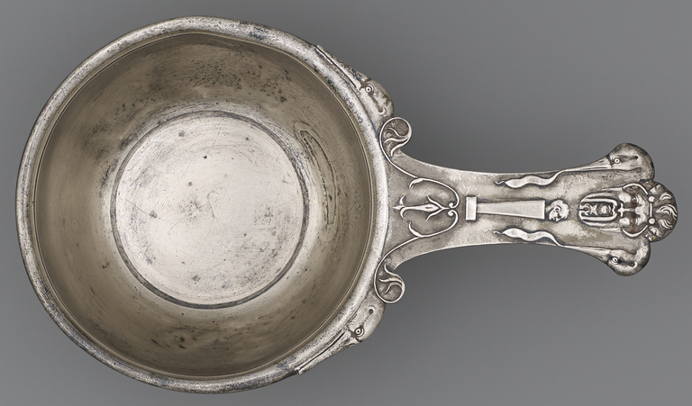 Photograph from above of a silver, long-handled bowl. The handle has a stylized, bearded male face, a male head in profile atop a pillar, and stylized bird heads and scrollwork.