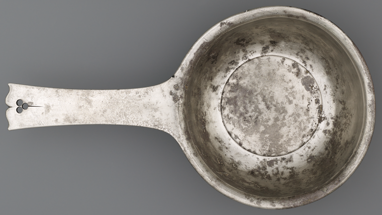 Photograph from above of a silver, long-handled bowl. The handle is devoid of decoration, with the exception of scrolled end and three holes arranged in a tight triangle.