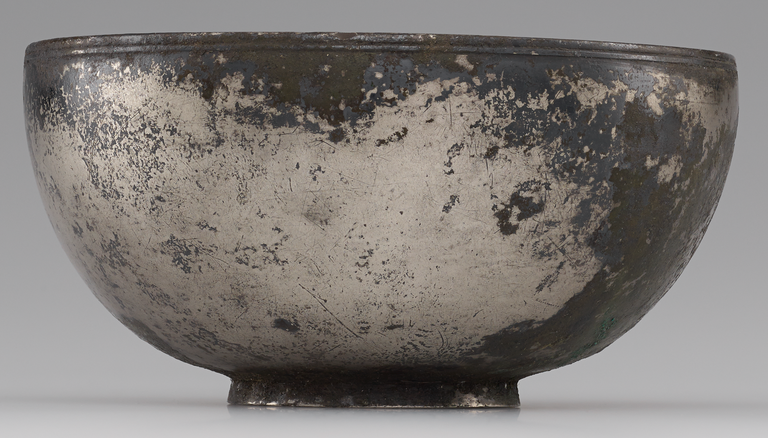 Photograph from the side of a hemispherical silver bowl with a short circular base and thin, banded rim. No figurative decoration is visible.