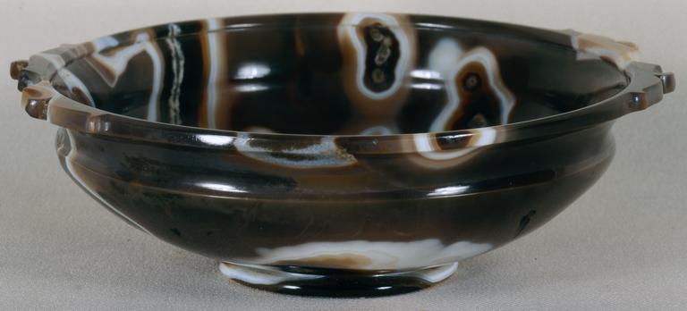 Photograph from the side and a bit above of a sardonyx bowl. A small, circular base supports the wide-flared body, which is topped by a narrow flat rim with projecting scalloped edges on two sides.