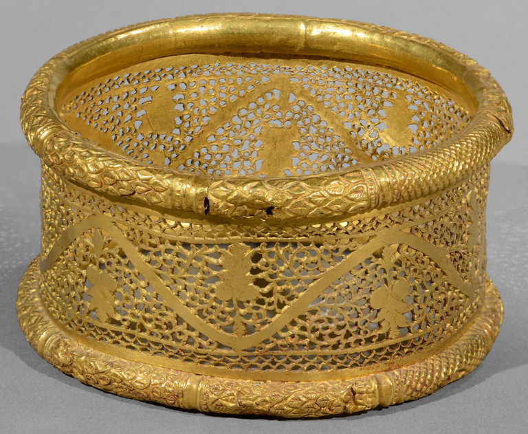 Photograph from the side of a cylindrical gold bracelet. The cylinder is worked in a see-through geometric pattern reminiscent of lace. A stylized grapevine with grapes runs around the cylinder. Both ends of the cylinder are closed with thick bands in circular cross section, patterned like foliage.