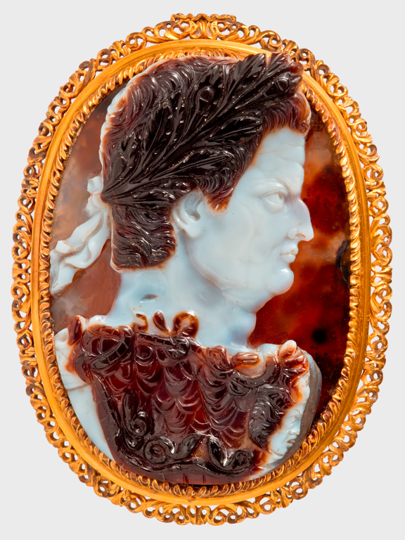 Photograph of oval cameo depicting a male figure in profile. The face and hair ribbons are white; the rest of the cameo a varied brownish red.  The cameo is set in a decorative gold frame.