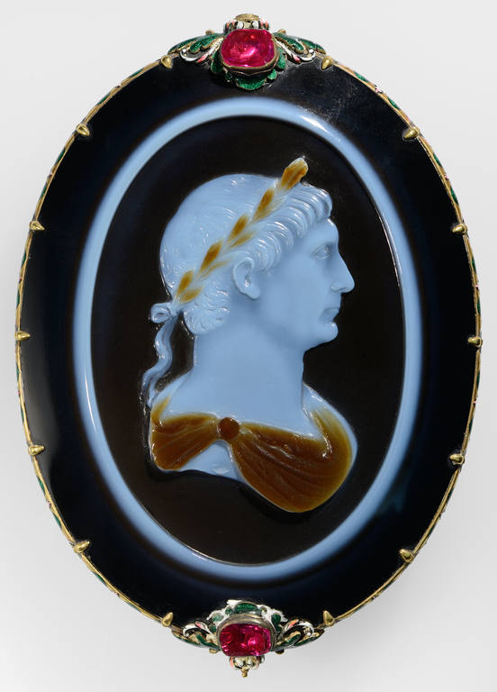 Photograph of oval cameo depicting a male figure in profile. The face and hair are white, as is an oval band surrounding the bust. The background of the cameo is black, but the shoulder drapery and leafy head band are mixed brown and white. The cameo is set in a decorative frame with set rubies at top and bottom.