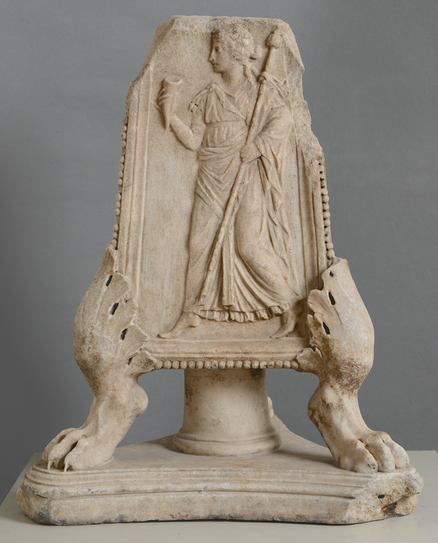 Photograph of a three-sided, white marble candelabra base resting on a short central pillar and three lion's feet. The side shown depicts a female figure in relief, striding and holding a staff in one hand and a horn in the other.