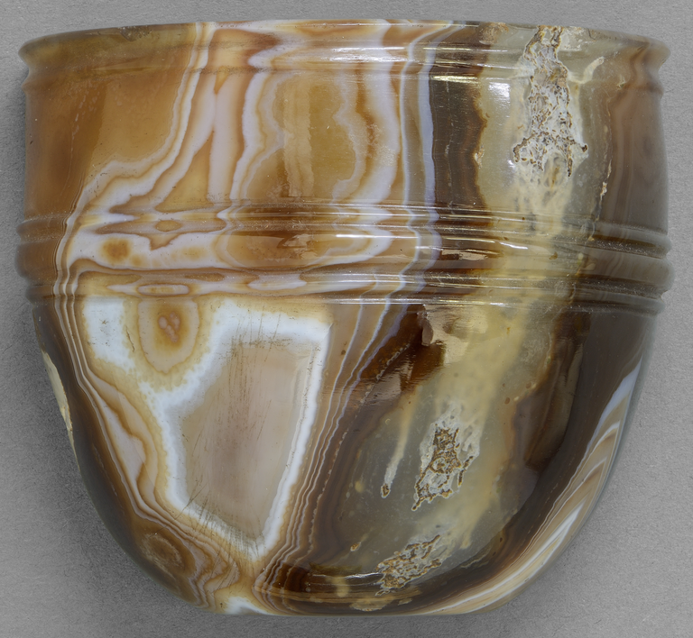 Photograph from the side of a sardonyx cup with a rounded base and a cylindrical upper body separated by a groved band. Another groved band the goes around the top.
