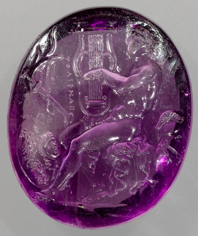 Photograph of an amethyst gem showing a nude male figure seated, playing a stringed instrument. Behind him foliage may be seen. At his feet lies a shield and to his left a sheathed sword, and bow. A Greek inscription runs vertically next to the cithara.
