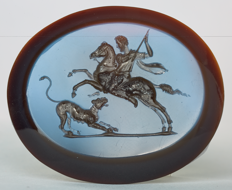 Photograph of an engraved gem depicting a leaping horse bearing a curly-haired and bearded rider with raised spear. Below and in front of a horse, a female tiger crouches.