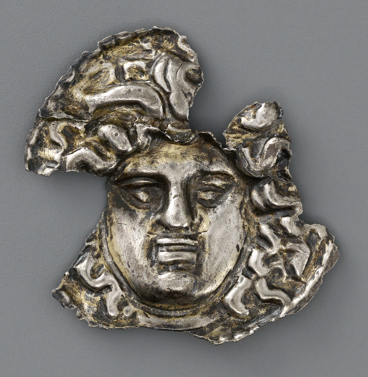 Photograph of a fragmentary silver and gold medallion featuring a face surrounded by a cloud of snakes.