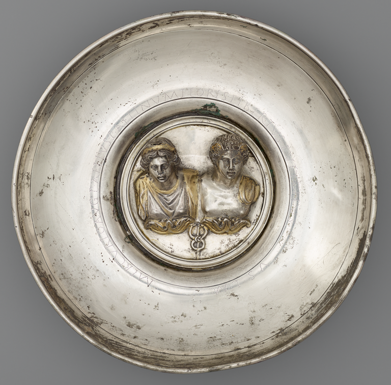 Photograph of a silver offering bowl with a large, central medallion in silver and gold depicting two busts: one male and one female. A caduceus appears below the busts.