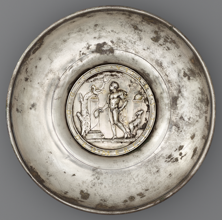 Photograph of a silver offering bowl with a large, central medallion depicting Mercury, nude with caduceus, standing before a tall pedastal or altar. A tree, a chicken, and a goat are also shown.