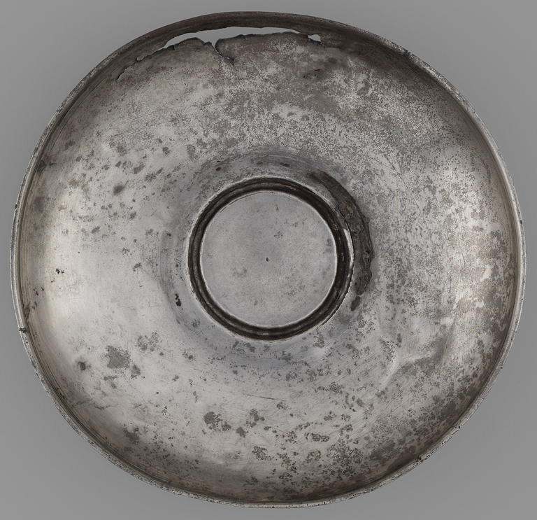 Photograph from above of a silver offering bowl. No figurative decoration is visible. Some corrosion and splitting apart of the bowl is visible.