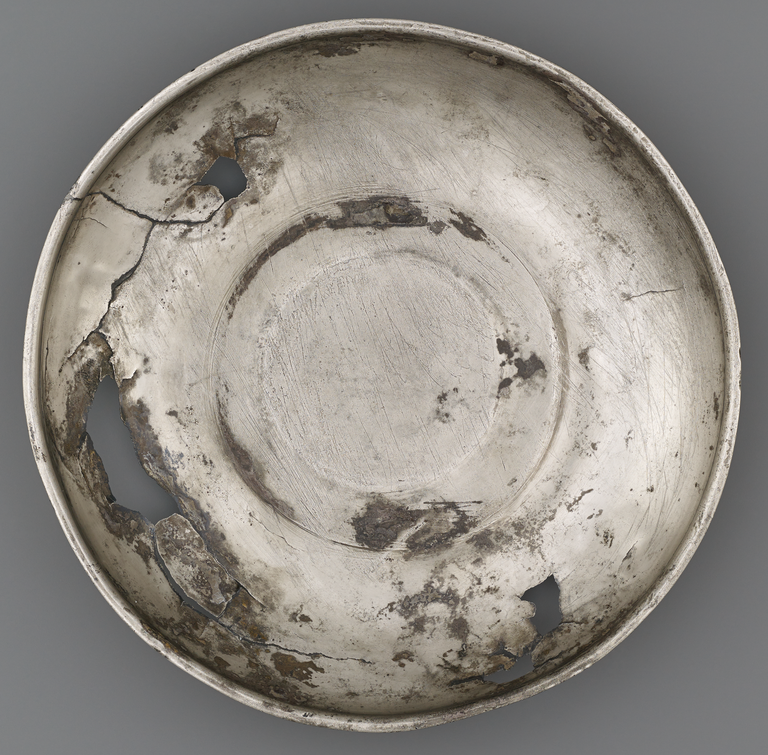Photograph from above of a silver offering bowl. No figurative decoration is visible. Cracks, holes, and some corrosion are visible.