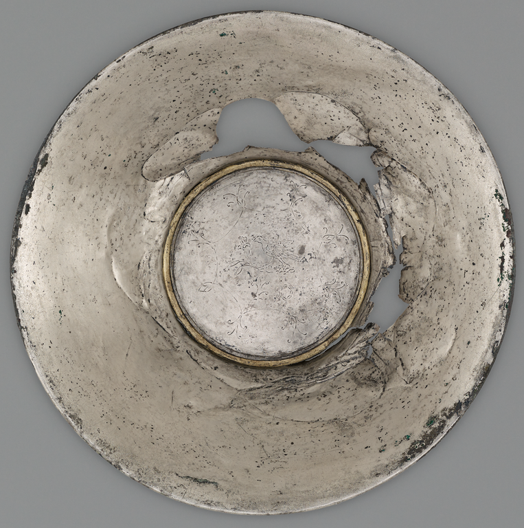 Photograph from above of a silver offering bowl. In the center is a flat medallion surrounded by a gold band. Incised on the medallion is a figure of a bird and stylized vegetation.