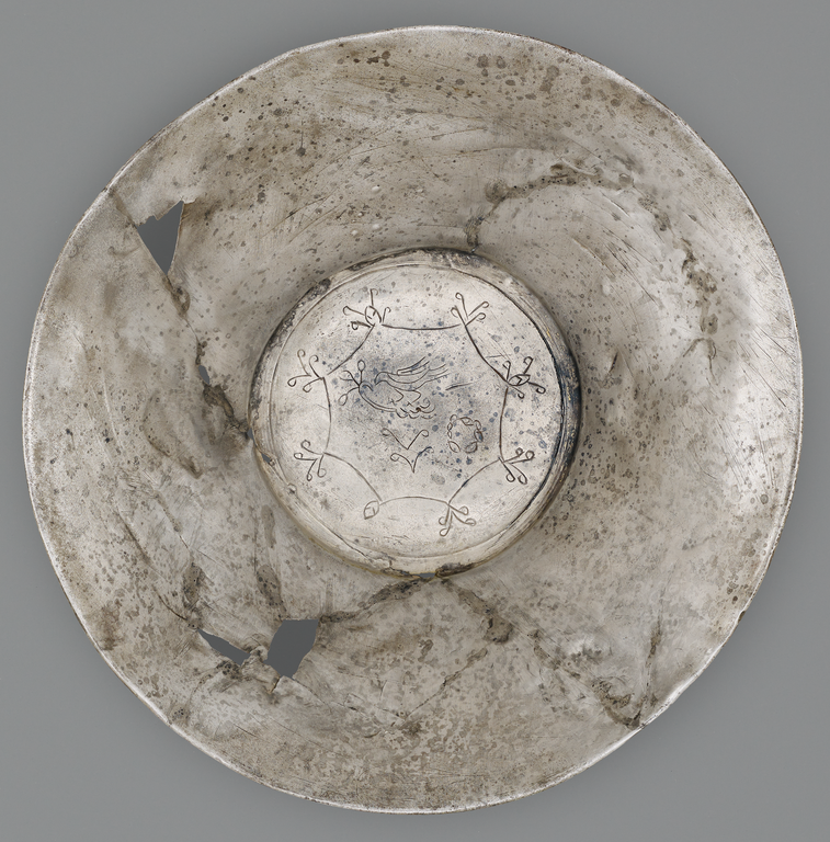 Photograph from above of a silver offering bowl. In the center is a flat medallion surrounded by a gold band. Incised on the medallion is a figure of a bird and stylized vegetation.