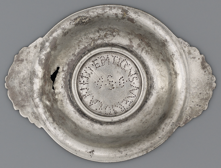 Photograph from above of a silver offering dish with two flat, rounded handles. A Latin inscription is incised in the circular center of the dish.