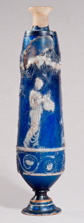 Photograph of a tall, blue perfume flask on which is depicted (in white cameo) a female figure holding a cornucopia. It is also decorated with stylized vegetation.