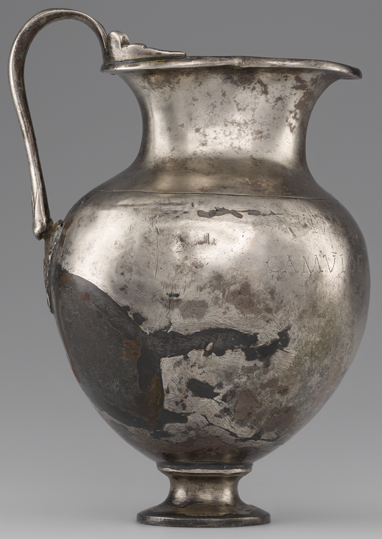 Photograph from the side of a silver pitcher with a single, looping handle. There is no visible decoration, but a Latin inscription is incised on the body of the pitcher on the spout side.