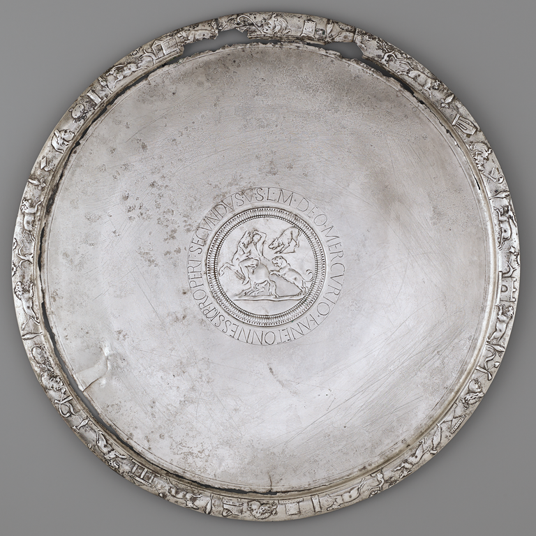 Photograph of a silver plate with a central medallion bearing a hunting scene, surrounded by an incised Latin text. The plate's rim is decorated with animals, buildings, and other objects.