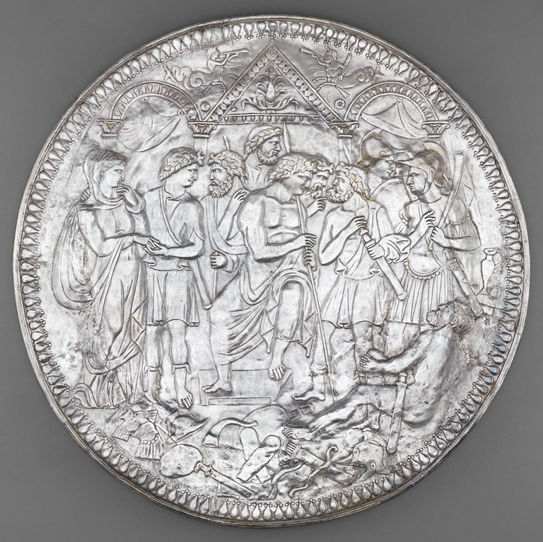 Photograph from above of a heavily illustrated, circular silver and gold plate depicting 8 male figures and 1 female figure (clothed) conferring in front of a stylized architectural facade. A naked man with a sword reclines at their feed; around him are pieces of armor and clothing.