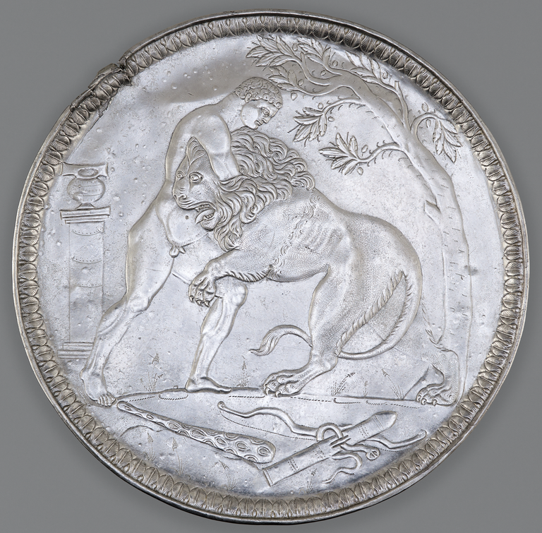 Photograph from above of a silver plate depicting Hercules wrestling the Nemean lion. A tree is depicted on the right and a round object atop a pillar appears on the left. Below the figures' feet are a club, a bow, and an empty quiver.