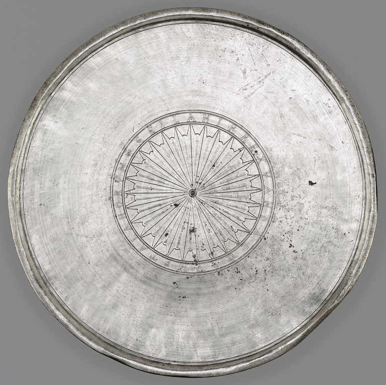 Photograph from above of a silver plate. A large medallion in the center bears an incised geometric startburst pattern. It is surrounded by an incised band in which a Latin inscription is visible.