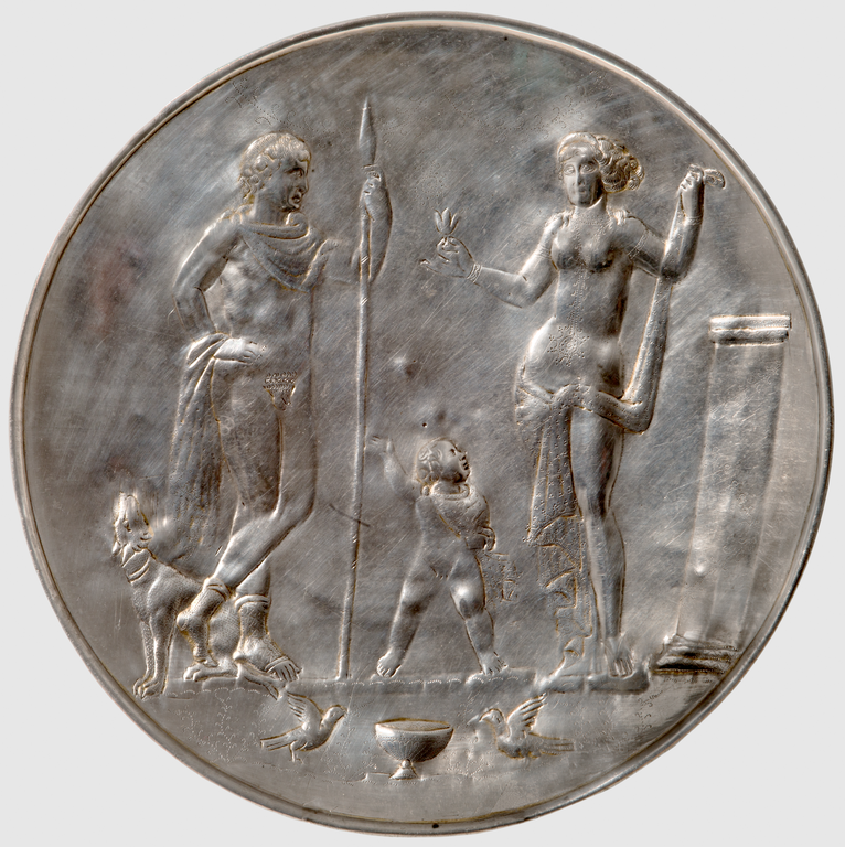 Photograph from above of a silver plate. At left, a nude male figure leans on his spear and gazes at a topless female figure at right holding leaves in one hand. A nude child also gazes at the female figure. At the man's foot sits a dog. Below the figures' feet, two birds flank a bowl. At right a pillar.