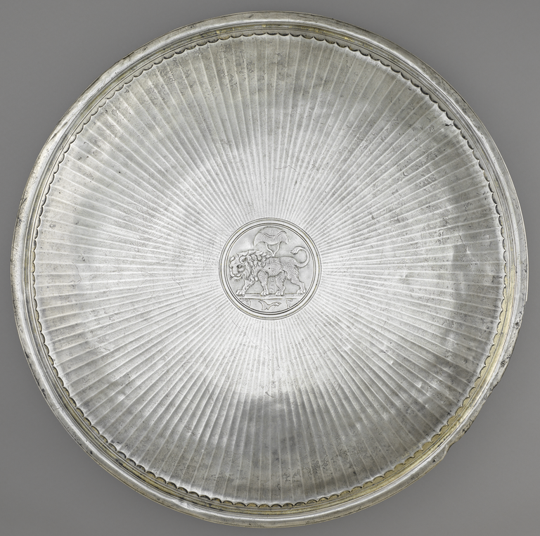 Photograph from above of a silver and gold plate. A fan pattern radiates from a central medallion in which is depicted a male lion in profile with a palm front above.