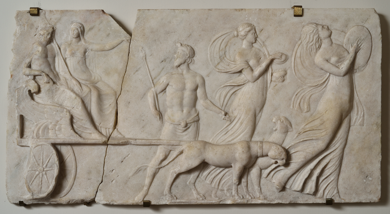 Photograph of a marble relief depicting a female figure holding a large, oval object, a second female figure with a lidded container, a bearded male figure with a thyrsus and a chariot, pulled by two panthers and carrying seated male and female figures.