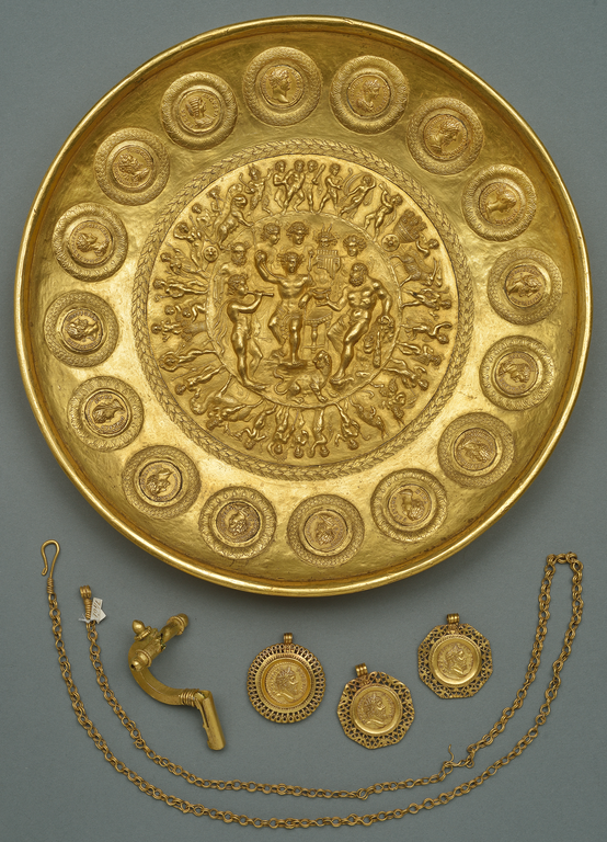 Photograph showing an ornate bowl, brooch, three coins in frames, and a chain, all made of gold.