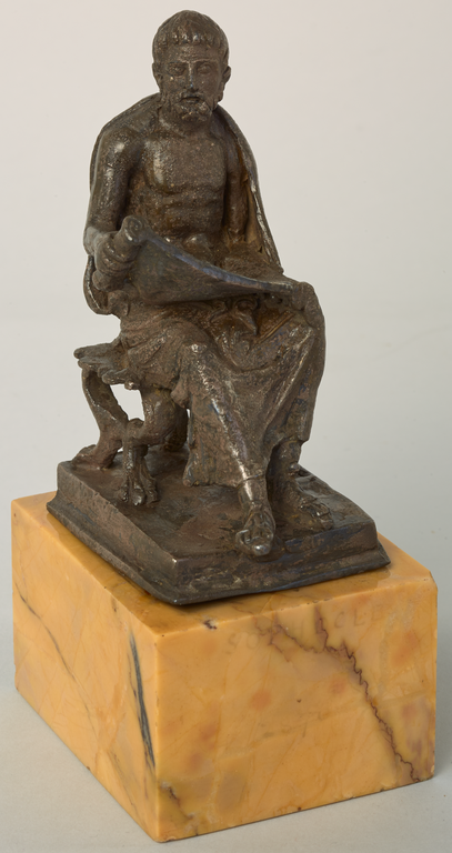 Photograph from the front of a silver statuette of a seated, bearded man looking down at a scroll that he holds open on his lap.