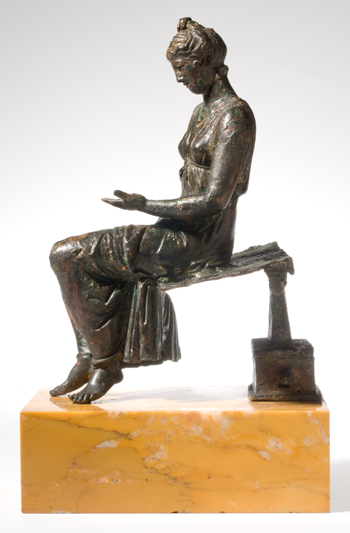 Photograph from the side of a bronze statuette depicting a seated young woman looking down at her hand, which is held horizontally above her lap.