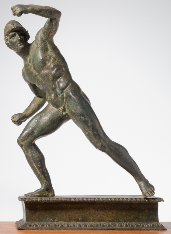 Photograph of a bronze statuette depicting a nude male figure wearing a helmet and striding forward. His head is turned to the side and he has raised one bent arm above his head.