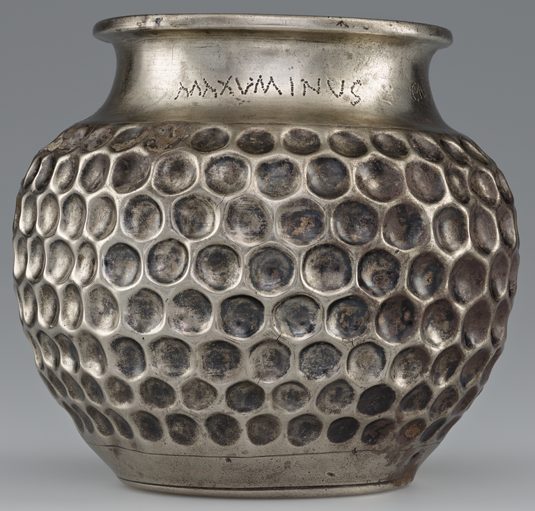 Photograph from the side of a silver vase whose body is decorated with a closely-spaced geometric pattern of circular indentations. A Latin inscription appears on the smooth neck, just below the rim.
