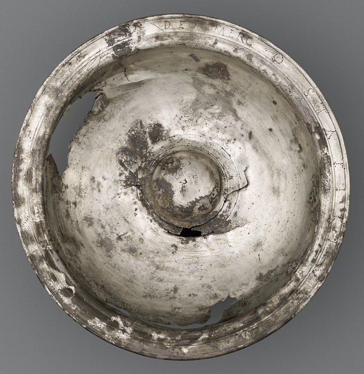 Photograph from above of a tarnished round bowl missing some of its body. On the wide flat rim is incised a Latin inscription.