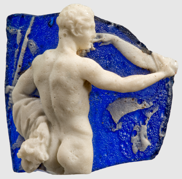 Photograph of a blue cameo glass fragment, depicting a nude male figure gesturing with one arm. The arm of another figure reaching back toward the male's mouth is also preserved.