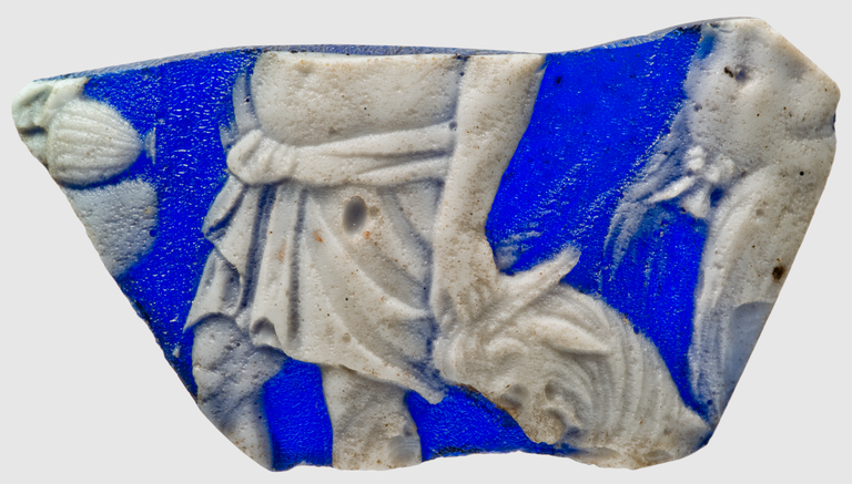 Photograph of a blue cameo glass fragment, depicting part of a male figure leading a ram. A second, nude male figure seems to have followed.